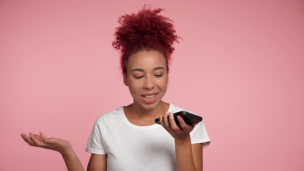 Close up smiling African American redheaded curly woman uses smartphone dictates an audio message to friend. Portrait positive happy female talking on mobile cell phone on isolated pink background - Footage, Video
