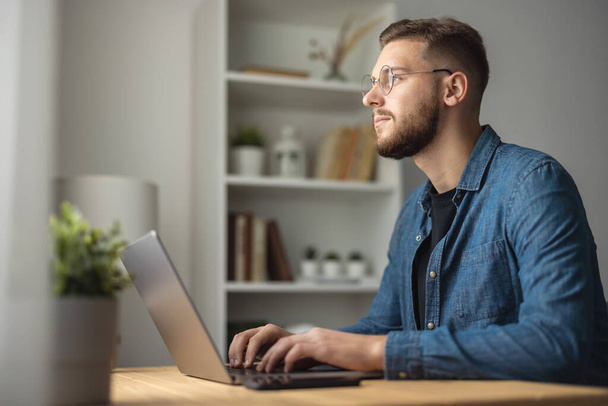 Bearded man in denim shirt working on laptop, coding on computer remotely, bright home interior - Φωτογραφία, εικόνα