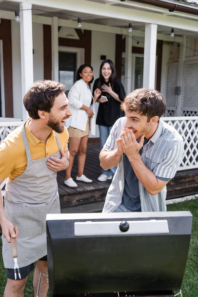 Веселые мужчины смеются возле гриля и размытые межрасовые друзья на заднем дворе  - Фото, изображение