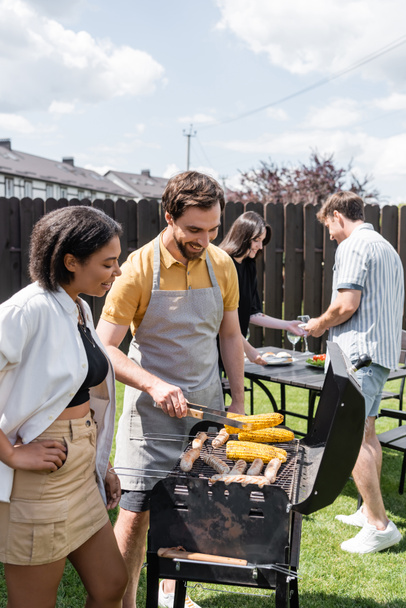 Alegre interracial amigos cocinar comida en la parrilla durante barbacoa en el patio trasero  - Foto, imagen