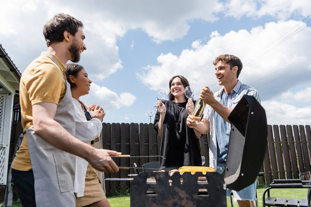 Uśmiechnięty mężczyzna i kobieta trzymający wino i kieliszki w pobliżu międzyrasowych przyjaciół i grilla na świeżym powietrzu  - Zdjęcie, obraz