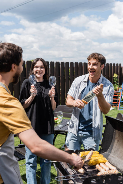 Cheerful man and woman holding wine and glasses near blurred friend cooking bbq in backyard  - Foto, immagini