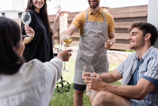 Uomo in grembiule versando vino vicino amici multietnici con occhiali all'aperto  - Foto, immagini