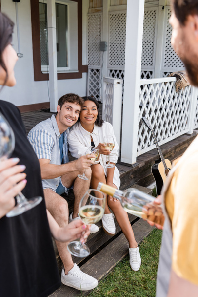 Smiling multiethnic friends holding wine near acoustic guitar and blurred people in backyard  - Foto, imagen