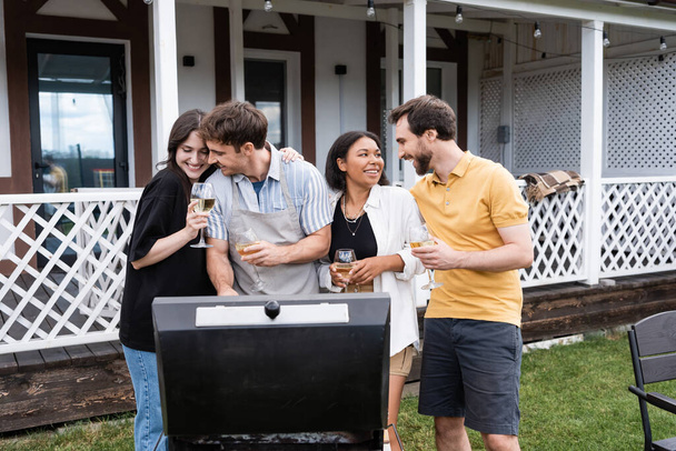 Glimlachende multi-etnische koppels met wijn praten in de buurt van grill in de achtertuin  - Foto, afbeelding