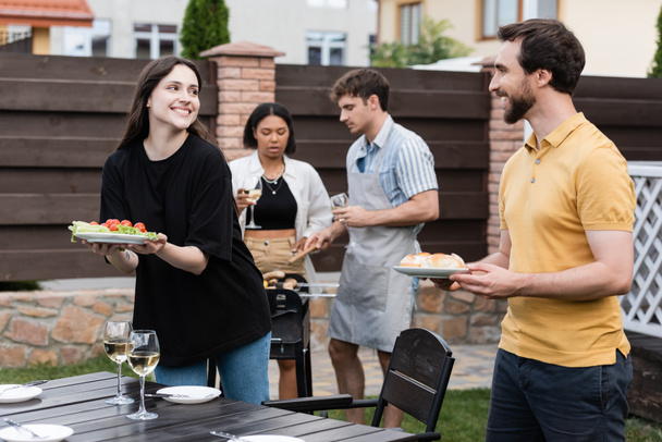 Positive people holding food near wine and interracial friends during picnic in backyard  - Foto, Imagem