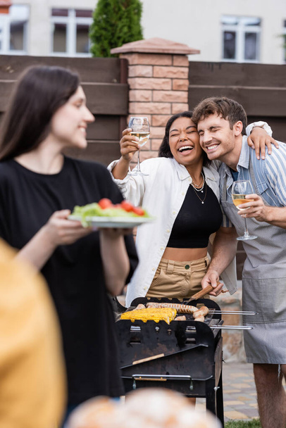 Heureuse femme bi-raciale tenant du vin et embrassant un ami dans un tablier faisant un barbecue à l'extérieur  - Photo, image
