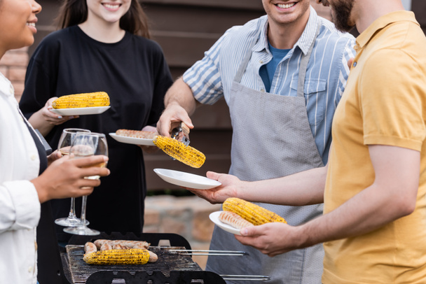 Przycięty widok uśmiechniętego mężczyzny w fartuchu trzymającego grillowaną kukurydzę w pobliżu międzyrasowych przyjaciół z talerzami i winem na świeżym powietrzu  - Zdjęcie, obraz