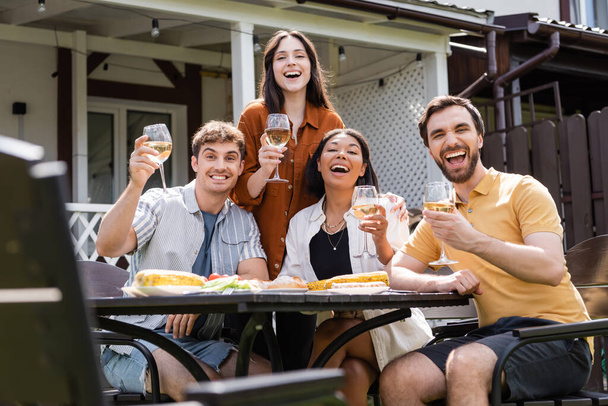 Happy multiethnic friends with wine looking at camera near food in backyard  - Photo, Image