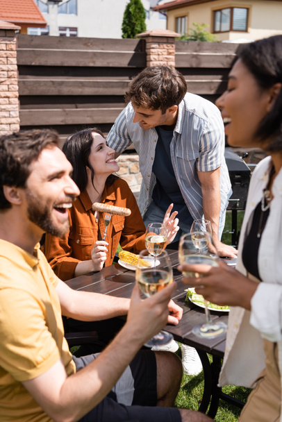 Smiling friends talking near food and blurred multiethnic people with wine in backyard  - Foto, Imagem