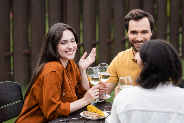 Smiling woman holding wine and talking to multiethnic friends near grilled food and smartphone outdoors  - Фото, изображение