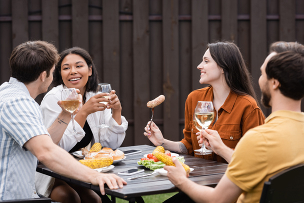 wesoły wielonarodowy przyjaciele trzymając kieliszki z winem i posiłek podczas imprezy bbq na podwórku  - Zdjęcie, obraz