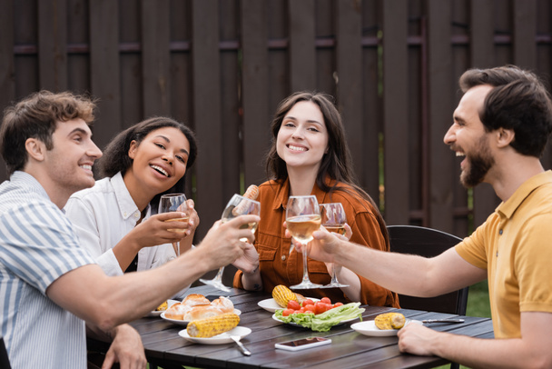 cheerful multiethnic friends clinking glasses with wine and having lunch during bbq party  - Фото, изображение