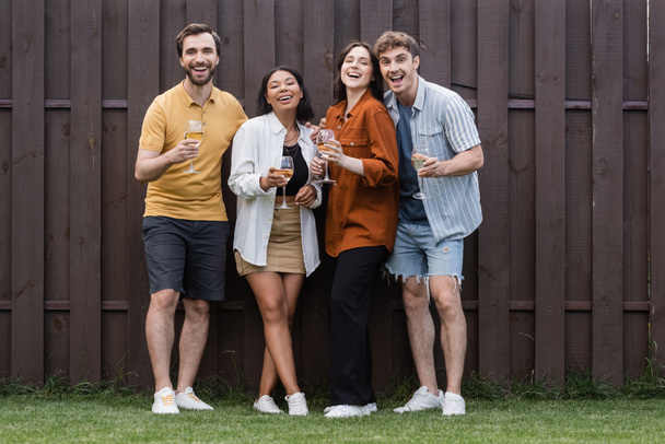 full length of happy interracial friends holding glasses with wine while standing near fence on backyard - Foto, Bild