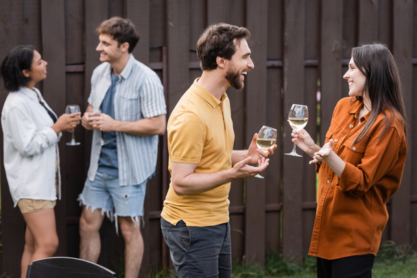 cheerful couple holding glasses with wine while talking near blurred interracial friends on blurred background - Foto, Imagem