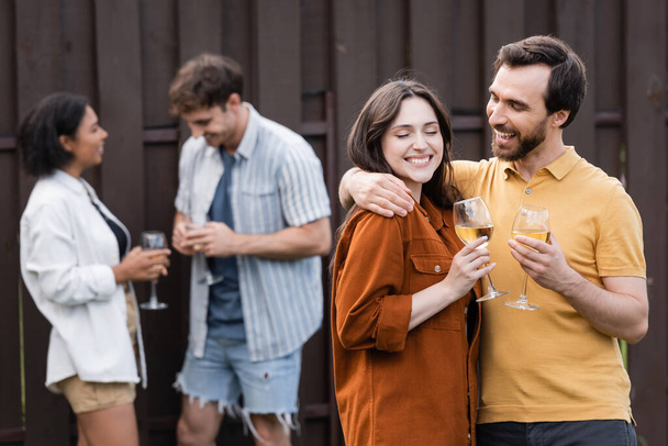 alegre pareja sosteniendo vasos con vino mientras abrazando cerca borrosa interracial amigos en borrosa fondo - Foto, imagen