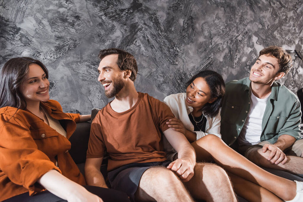amis multiculturels positifs en vêtements décontractés assis sur le canapé gris dans le salon moderne - Photo, image