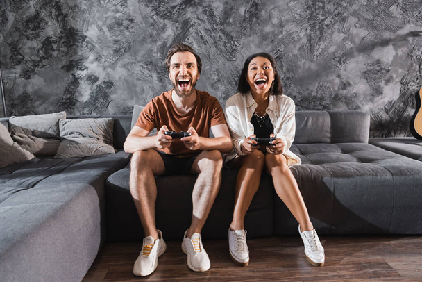 KYIV, UKRAINE - JULY 26, 2022: amazed interracial friends playing video game and sitting on grey couch - Fotografie, Obrázek