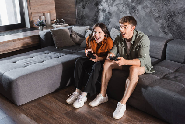 KYIV, UKRAINE - JULY 26, 2022: excited friends holding joysticks and playing video game while sitting on grey couch - Foto, Imagen