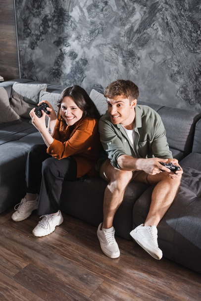 KYIV, UKRAINE - JULY 26, 2022: cheerful friends holding joysticks and playing video game while sitting on grey couch - Fotó, kép