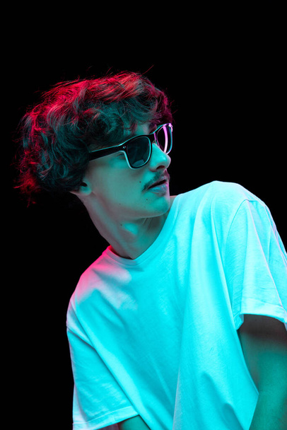 Ein junger, stylischer Mann mit Schnurrbart posiert mit Sonnenbrille und weißem T-Shirt vor schwarzem Hintergrund in Neonlicht. Konzept der Jugend, Mode, Lifestyle, Emotionen, Mimik. Kopierraum für Werbung - Foto, Bild