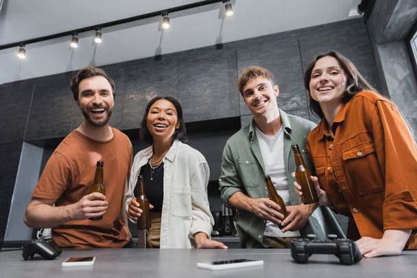 KYIV, UCRAINA - 26 LUGLIO 2022: amici multietnici sorridenti che tengono bottiglie di birra vicino a joystick e smartphone sul piano di lavoro della cucina - Foto, immagini