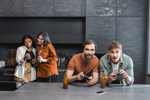 KYIV, UCRAINA - 26 LUGLIO 2022: uomini felici che giocano ai videogiochi vicino a donne interrazziali sfocate usando smartphone in cucina  - Foto, immagini