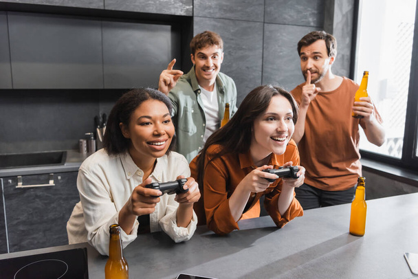 KYIV, UKRAINE - JULY 26, 2022: happy interracial women playing video game near man with beer showing hush sign in kitchen  - Foto, Bild