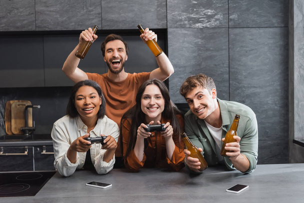 KYIV, UKRAINE - JULY 26, 2022: happy interracial women playing video game near cheerful male friends with beer in kitchen  - Фото, изображение