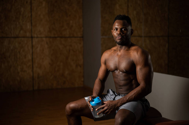 Hemdloser afrikanisch-amerikanischer Mann, der im Fitnessstudio aus einem Shaker trinkt - Foto, Bild