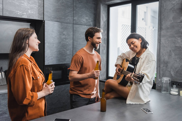 счастливая двурасовая женщина играет на акустической гитаре рядом с друзьями с бутылками пива - Фото, изображение