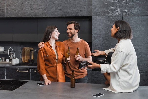 happy man hugging woman near bi-racial friend with acoustic guitar gesturing in kitchen - Photo, Image