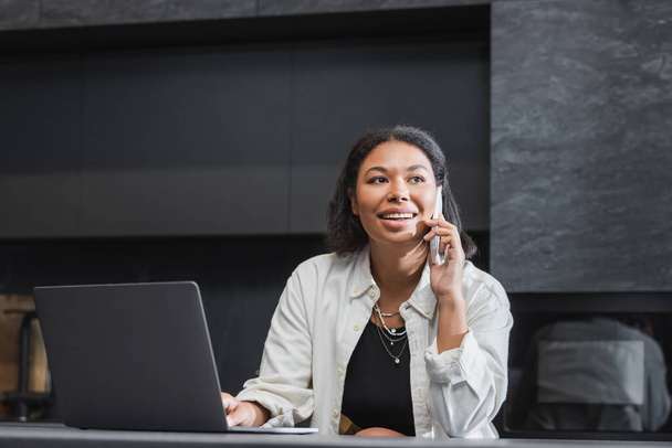 šťastná bi-rasová žena mluví na smartphone v blízkosti notebooku na pracovní desce  - Fotografie, Obrázek