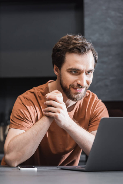 cheerful bearded freelancer looking at laptop near smartphone on kitchen worktop - Фото, зображення