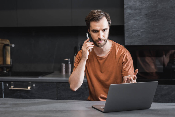 serious bearded freelancer talking on smartphone and looking at laptop  - Photo, image