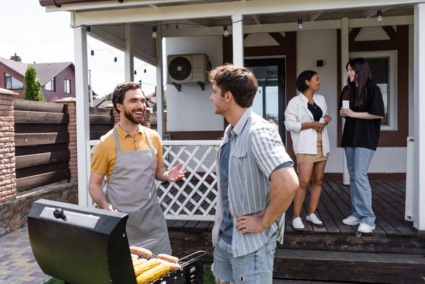 alegres hombres haciendo barbacoa y hablando cerca borrosa interracial amigos en el patio trasero  - Foto, Imagen