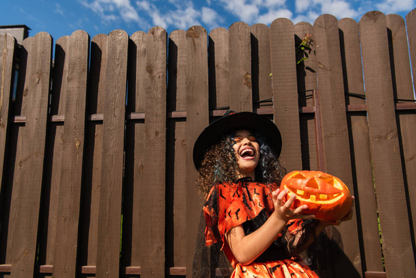 excited girl in witch halloween costume holding jack o lantern and laughing near wooden fence - Zdjęcie, obraz