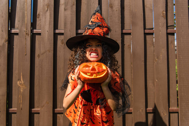 dívka v čarodějnice kostým drží jack o lucerna a ukazuje děsivý grimasa  - Fotografie, Obrázek