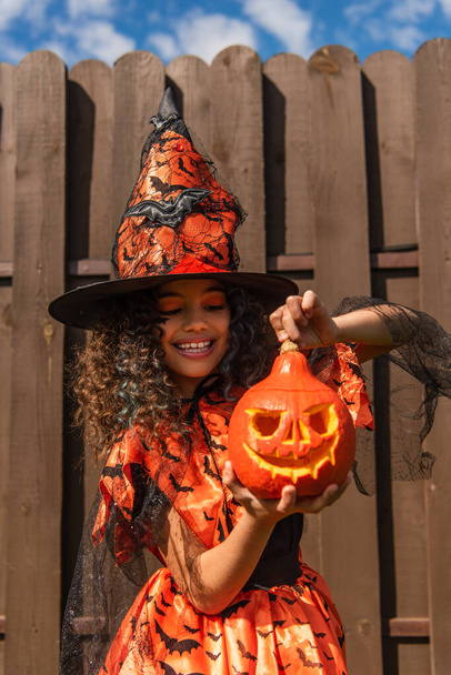 happy curly girl in halloween costume and witch hat holding carved pumpkin - Фото, изображение