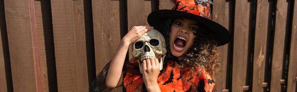 dívka v kostýmu čarodějnice drží lebku a vrčí s hněvivým úšklebkem, prapor - Fotografie, Obrázek