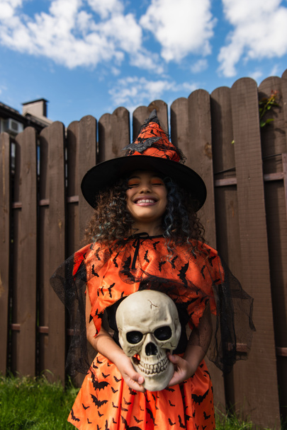 smiling girl in witch costume and pointed hat standing with closed eyes and scary skull - Fotografie, Obrázek