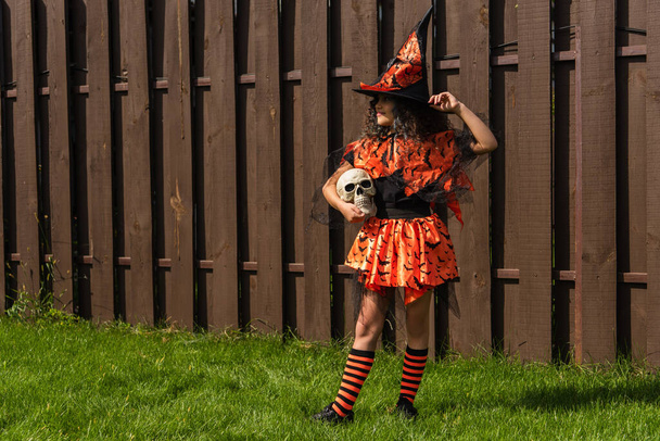 Mädchen in voller Länge im Halloween-Kostüm und gestreiften Kniestrümpfen mit Totenkopf am Holzzaun - Foto, Bild