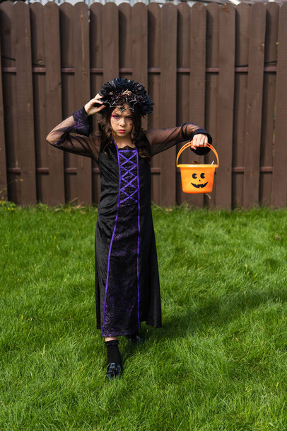 serious girl in black dress and wreath holding halloween bucket while walking in backyard - Foto, Imagen