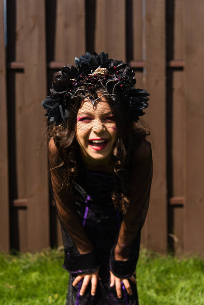 cheerful girl in halloween costume and black wreath laughing at camera outdoors - Photo, image