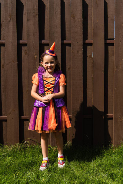 full length of girl in clown costume and party cap smiling near wooden fence - Fotoğraf, Görsel