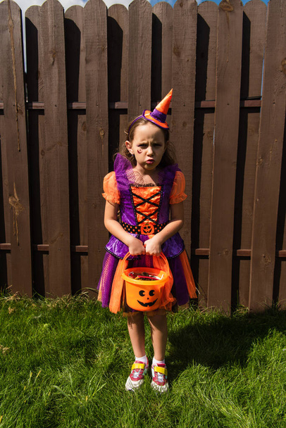 offended girl in clown costume holding halloween bucket with sweets and looking at camera - Foto, Bild