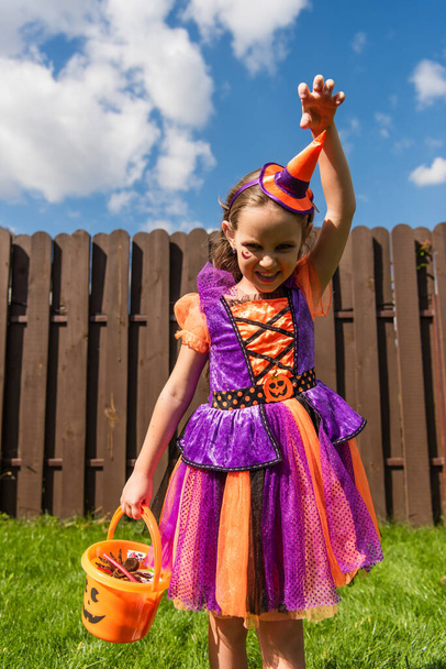 girl in clown costume showing scary grimace and frightening gesture while holding halloween bucket - Foto, Imagen