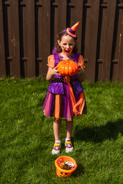 menina emocionada em traje de palhaço segurando abóbora perto de balde de Halloween com doces - Foto, Imagem