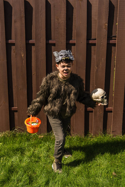 longitud completa de asiático chico en hombre lobo traje corriendo con cráneo y halloween cubo con dulces en patio trasero - Foto, Imagen