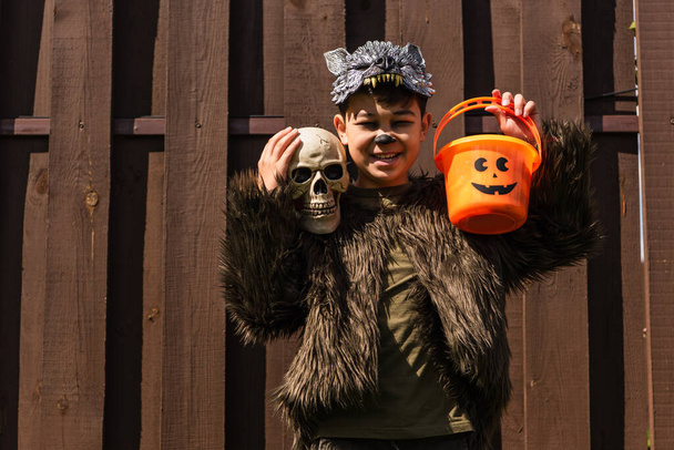 wesoły azjatycki chłopiec w wilkołak kostium gospodarstwa czaszki i wiadro halloween - Zdjęcie, obraz